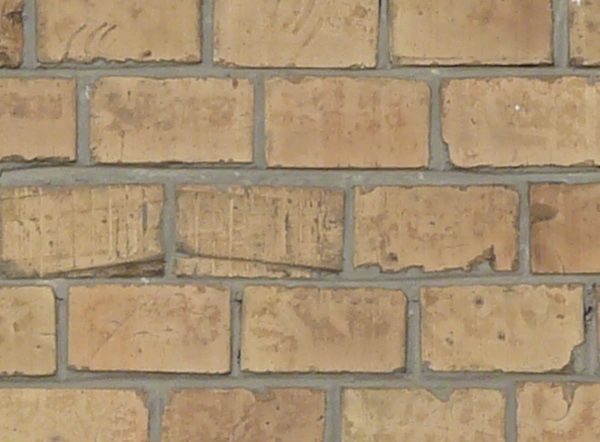 Brick fence texture made up of bricks in various shades of tan and brown, with a strip of grey cement with brick fragments near the bottom.